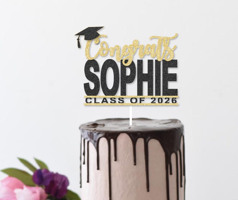 May include: A cake topper with the words "Congrats Sophie" in gold glitter script and black block letters, topped with a graduation cap. The text "Class of 2026" is also included. The topper is placed on a cake with chocolate frosting.