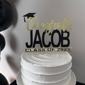 May include: A white frosted cake with a decorative topper that reads "Congrats JACOB CLASS OF 2023". The topper is black with gold glitter accents and a graduation cap. Black balloons are visible in the background.