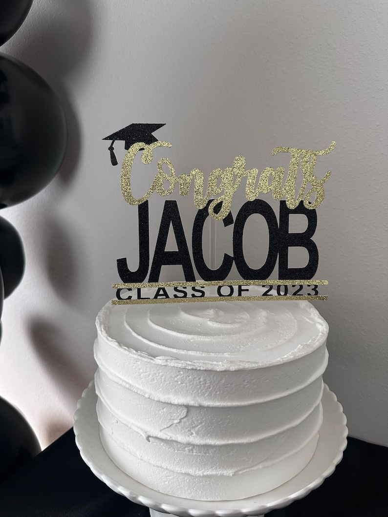 May include: A white frosted cake with a decorative topper that reads "Congrats JACOB CLASS OF 2023". The topper is black with gold glitter accents and a graduation cap. Black balloons are visible in the background.