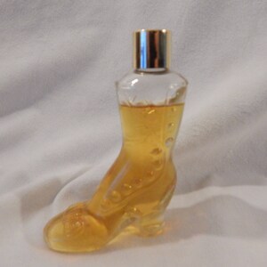 May include: A clear glass bottle shaped like a shoe, filled with a light yellow liquid. The bottle has a gold-colored cap.