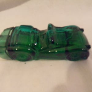 May include: A green glass car shaped bottle. The bottle is a vintage style car with a rounded body and a single driver's seat.
