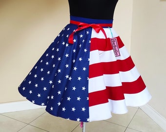 May include: A red, white, and blue circle skirt with a black top and a red ribbon. The skirt is made of fabric with a stars and stripes pattern. The skirt has a tag that says "Be Bold".