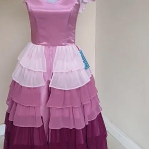 May include: A layered, floor-length dress in shades of pink and purple, displayed on a mannequin. The bodice is a solid pink with a V-neck, and the skirt features multiple tiers of ruffled fabric. The dress has short, ruffled sleeves and a tag that says "Be Bouncy".