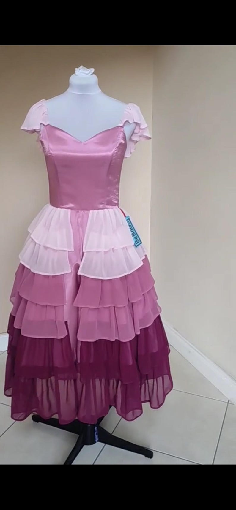 May include: A layered, floor-length dress in shades of pink and purple, displayed on a mannequin. The bodice is a solid pink with a V-neck, and the skirt features multiple tiers of ruffled fabric. The dress has short, ruffled sleeves and a tag that says "Be Bouncy".
