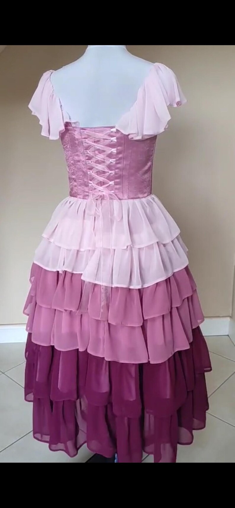 May include: A layered, floor-length dress in shades of pink and purple. The bodice is a deep pink with a lace-up back. The skirt features multiple tiers of ruffled fabric, transitioning from light pink to deep purple.