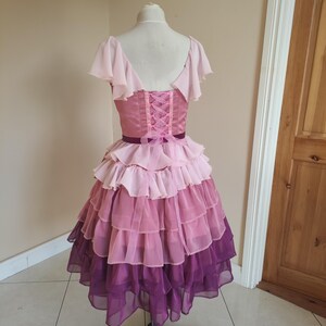May include: A layered, pink and purple dress on a mannequin. The bodice is a solid pink with a corset-style back. The skirt features multiple layers of ruffled fabric, transitioning from light pink to deep purple. The dress has short, flutter sleeves.