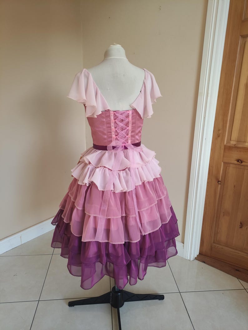 May include: A layered, pink and purple dress on a mannequin. The bodice is a solid pink with a corset-style back. The skirt features multiple layers of ruffled fabric, transitioning from light pink to deep purple. The dress has short, flutter sleeves.
