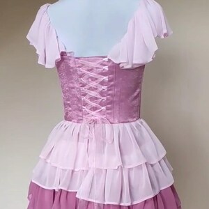May include: A lilac-colored dress with a corset back and layered skirt. The bodice features a satin-like fabric with a lace-up design. The skirt has multiple tiers of ruffled fabric in shades of pink and lavender. The dress has short, ruffled sleeves.