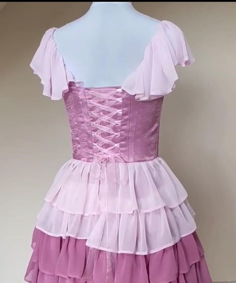 May include: A lilac-colored dress with a corset back and layered skirt. The bodice features a satin-like fabric with a lace-up design. The skirt has multiple tiers of ruffled fabric in shades of pink and lavender. The dress has short, ruffled sleeves.