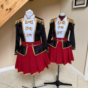 Puede incluir: Dos uniformes de gala de terciopelo rojo y negro con ribetes dorados y lazos dorados. Los uniformes están en maniquíes y tienen un cuello blanco y un cinturón negro con ribetes dorados.