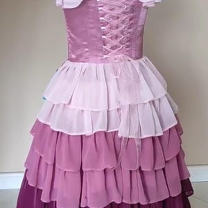 May include: A layered, ombre-effect dress in shades of pink and purple. The bodice features a corset-style back with ribbon lacing. The dress has ruffled sleeves and a tiered skirt, creating a flowing silhouette. The dress is displayed on a mannequin.