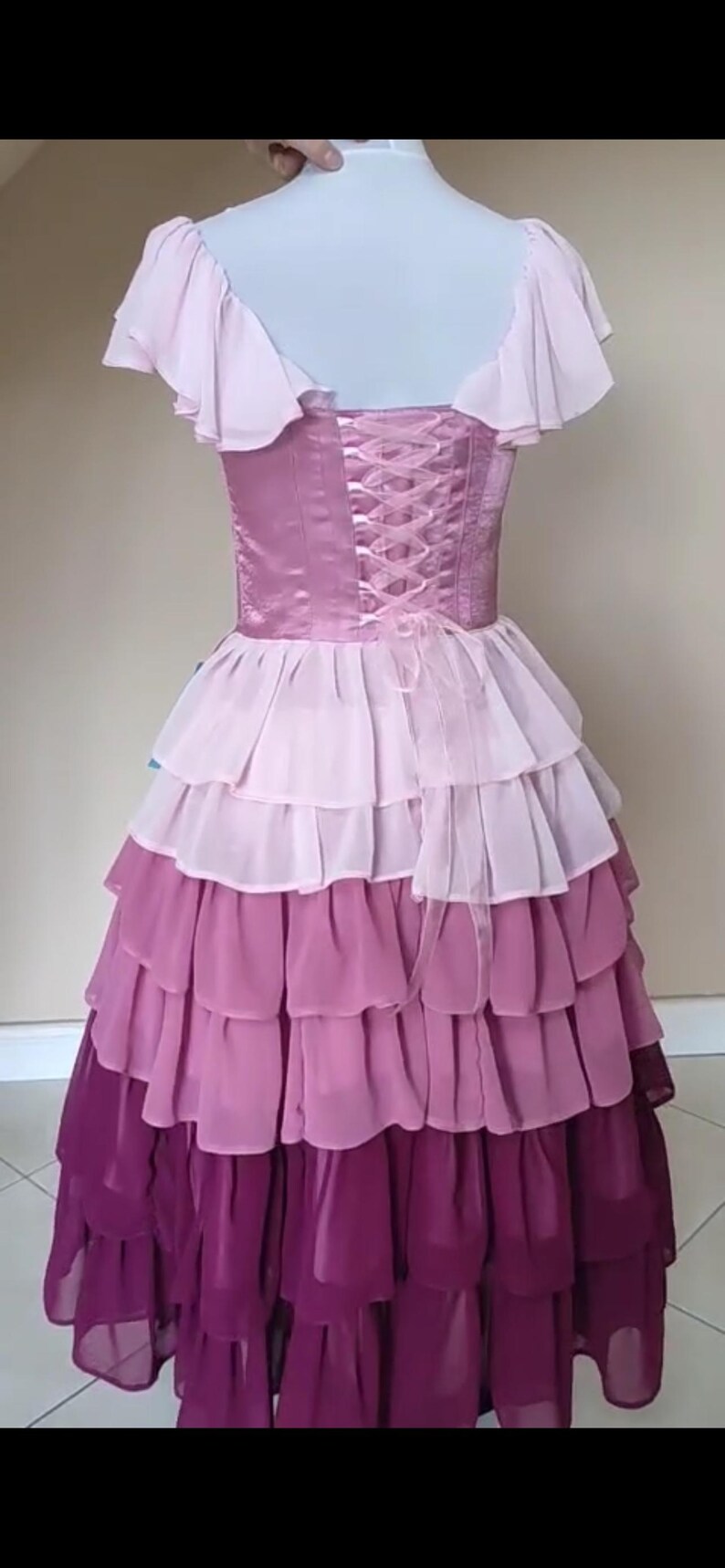 May include: A layered, ombre-effect dress in shades of pink and purple. The bodice features a corset-style back with ribbon lacing. The dress has ruffled sleeves and a tiered skirt, creating a flowing silhouette. The dress is displayed on a mannequin.