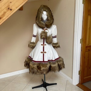 Peut inclure: Un manteau d'hiver blanc avec une capuche et une garniture en fourrure, présenté sur un mannequin. Le manteau présente des accents de fourrure marron, une bordure bordeaux et des fermetures décoratives. La robe a une jupe évasée et des manches longues.