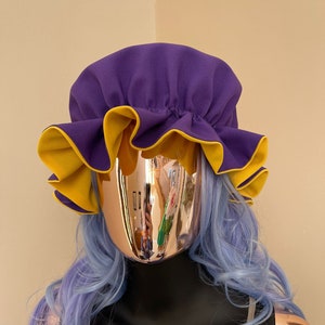Può includere: Un cappello di stoffa con volant viola e giallo. Il cappello è realizzato in un materiale morbido ed elastico e ha un elastico arricciato sul retro.