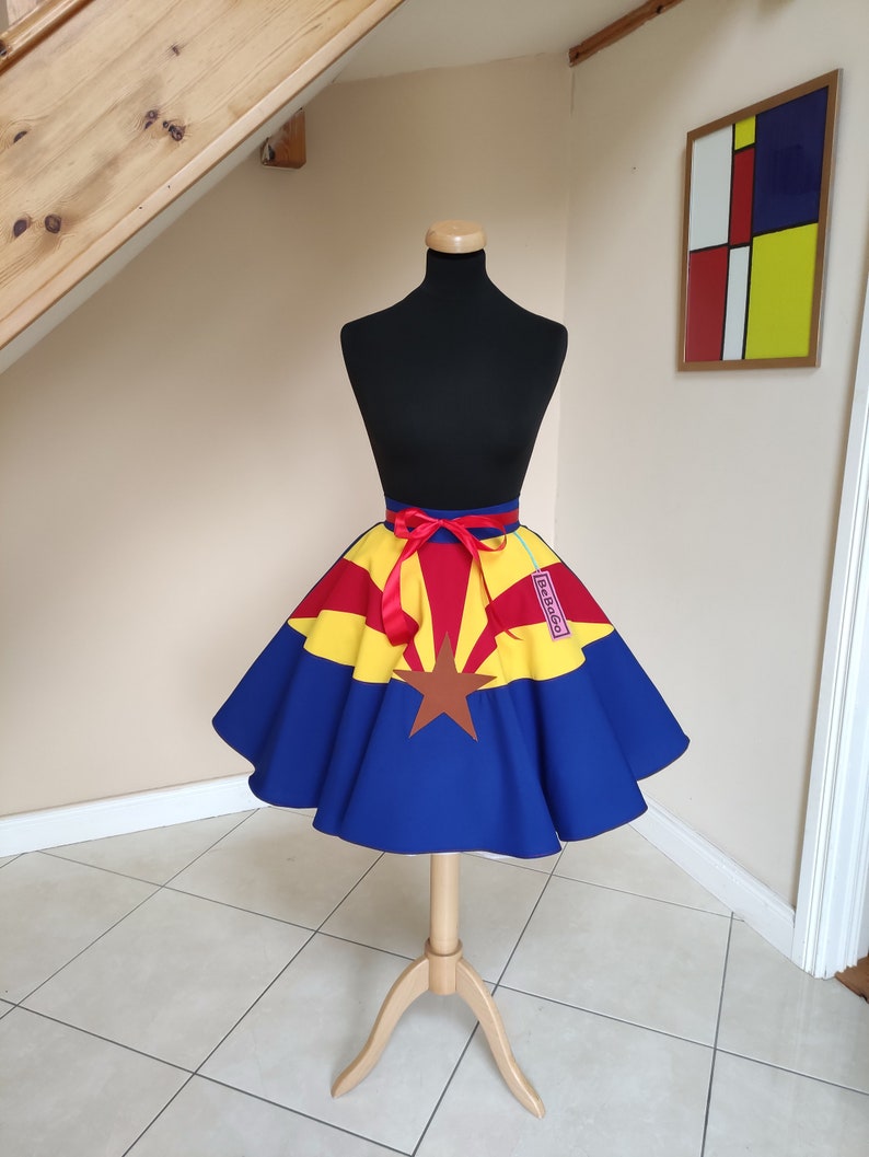 May include: A blue, yellow and red circle skirt with a star and the state of Arizona printed on it. The skirt is on a mannequin and has a red ribbon tied around the waist.
