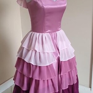May include: A layered, floor-length dress in shades of pink and purple. The bodice is a solid, shiny pink with a sweetheart neckline and cap sleeves. The skirt features multiple layers of ruffled fabric, transitioning from light pink to deep purple.
