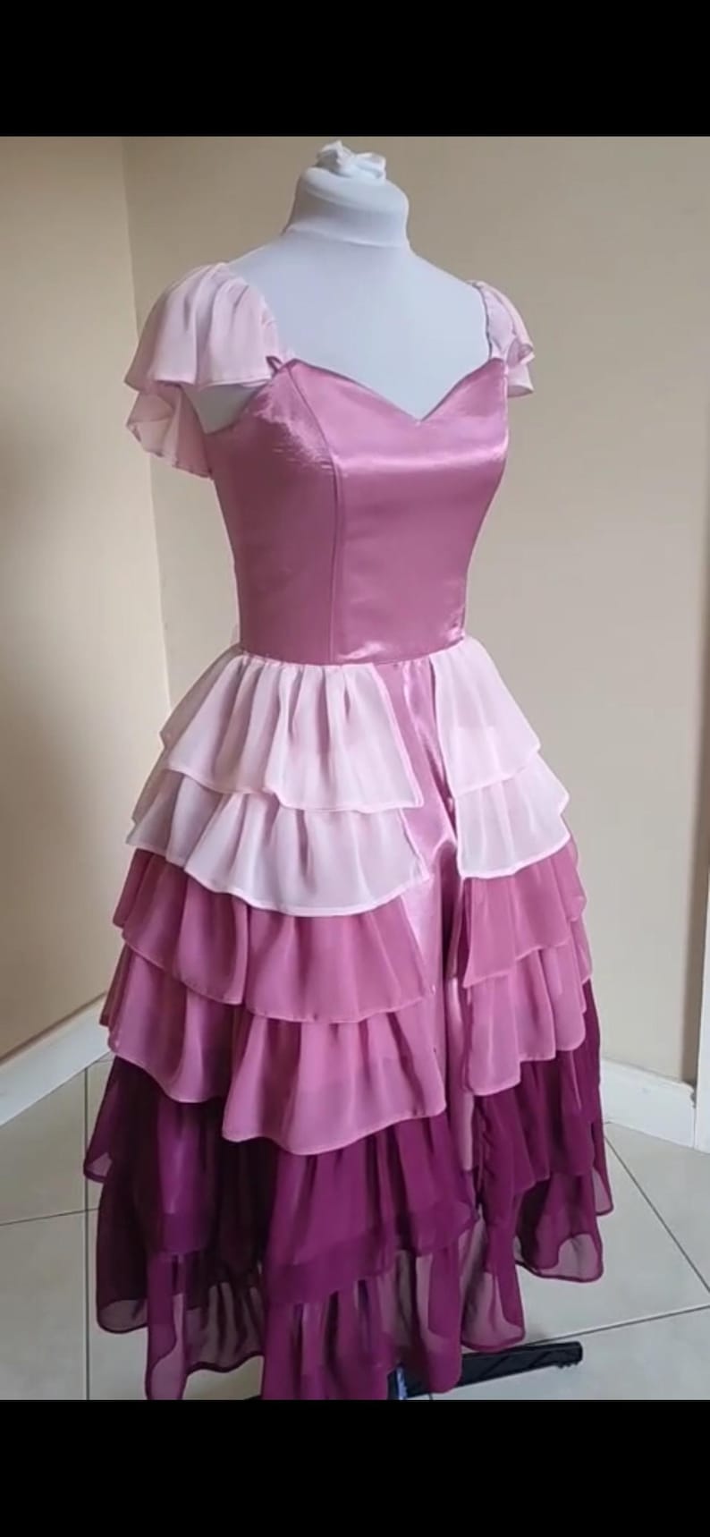 May include: A layered, floor-length dress in shades of pink and purple. The bodice is a solid, shiny pink with a sweetheart neckline and cap sleeves. The skirt features multiple layers of ruffled fabric, transitioning from light pink to deep purple.