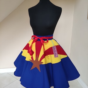 May include: A blue, yellow and red circle skirt with the Arizona state flag design. The skirt has a red ribbon tied around the waist and a pink tag that says "BeBago".