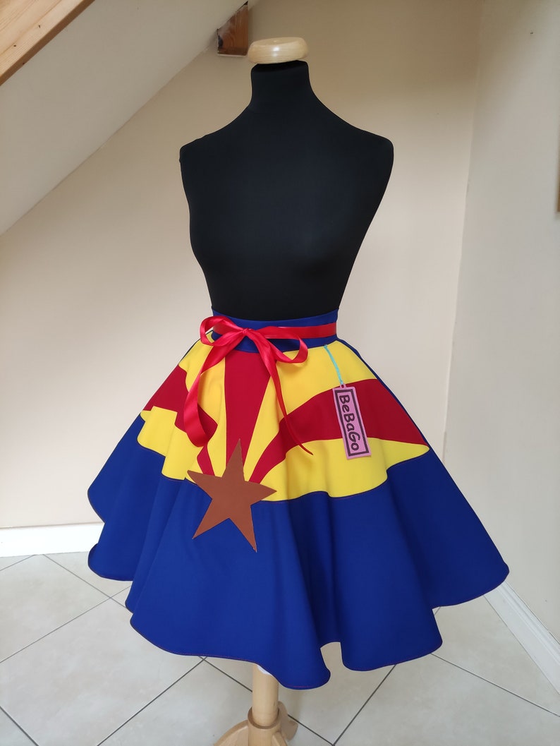 May include: A blue, yellow and red circle skirt with the Arizona state flag design. The skirt has a red ribbon tied around the waist and a pink tag that says "BeBago".