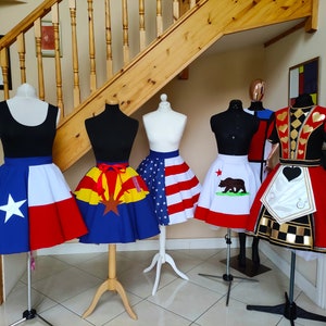 May include: Five different skirts on mannequins, each with a different state flag design. The skirts are all red, white and blue, with a variety of designs. The skirts are all on display in a room with a wooden staircase in the background.