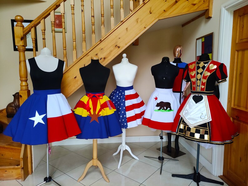 May include: Five different skirts on mannequins, each with a different state flag design. The skirts are all red, white and blue, with a variety of designs. The skirts are all on display in a room with a wooden staircase in the background.