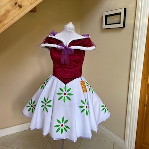 Puede incluir: Vestido burdeos y blanco con estampado floral. El corpiño es burdeos y la falda es blanca con diseños de flores verdes y rojas. El vestido tiene cuello blanco y detalles morados. Una etiqueta está sujeta a la falda.