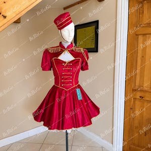 May include: A red dress with short sleeves, a high collar, and gold trim, featuring a front lace-up design with gold buttons. The outfit includes a matching red pillbox hat with gold accents. A small turquoise tag with the text BeBaGo is attached.