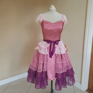 May include: A pink and purple dress with a sweetheart neckline and layered skirt. The bodice is a solid pink, and the skirt features multiple layers of ruffled fabric in shades of pink and purple. A purple ribbon belt cinches the waist.