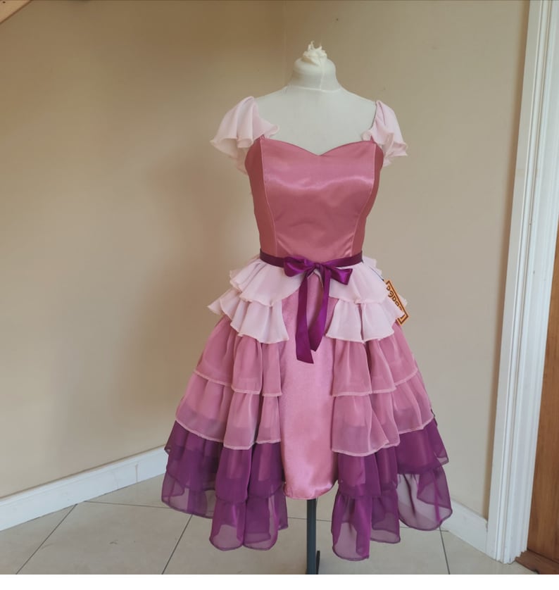 May include: A pink and purple dress with a sweetheart neckline and layered skirt. The bodice is a solid pink, and the skirt features multiple layers of ruffled fabric in shades of pink and purple. A purple ribbon belt cinches the waist.
