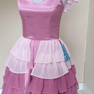 May include: A layered, pink and purple dress displayed on a mannequin. The bodice is a solid, satin-like material, while the skirt features multiple layers of ruffled fabric in varying shades of pink and purple. The dress has a V-neckline and short, ruffled sleeves.