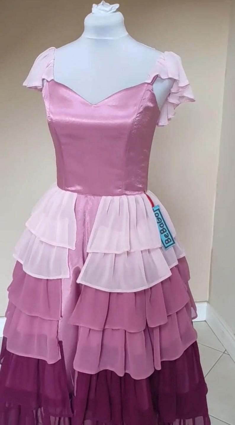 May include: A layered, pink and purple dress displayed on a mannequin. The bodice is a solid, satin-like material, while the skirt features multiple layers of ruffled fabric in varying shades of pink and purple. The dress has a V-neckline and short, ruffled sleeves.