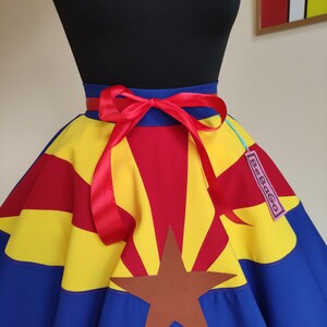 May include: A blue, yellow and red circle skirt with a brown star and a red ribbon tied at the waist. The skirt is printed with the Arizona state flag. The skirt has a tag that says "SeeBaGo".