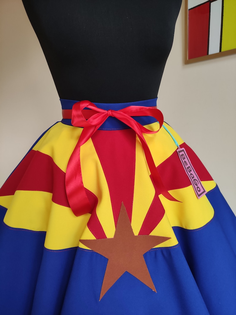 May include: A blue, yellow and red circle skirt with a brown star and a red ribbon tied at the waist. The skirt is printed with the Arizona state flag. The skirt has a tag that says "SeeBaGo".