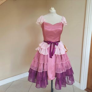May include: A vintage-style dress in shades of pink and purple, displayed on a mannequin. The dress features a satin bodice, ruffled skirt, and a purple ribbon belt. The skirt has multiple layers of ruffles, transitioning from light pink to deep purple.