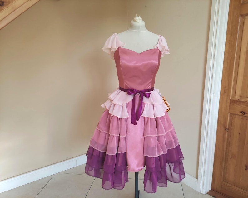 May include: A vintage-style dress in shades of pink and purple, displayed on a mannequin. The dress features a satin bodice, ruffled skirt, and a purple ribbon belt. The skirt has multiple layers of ruffles, transitioning from light pink to deep purple.