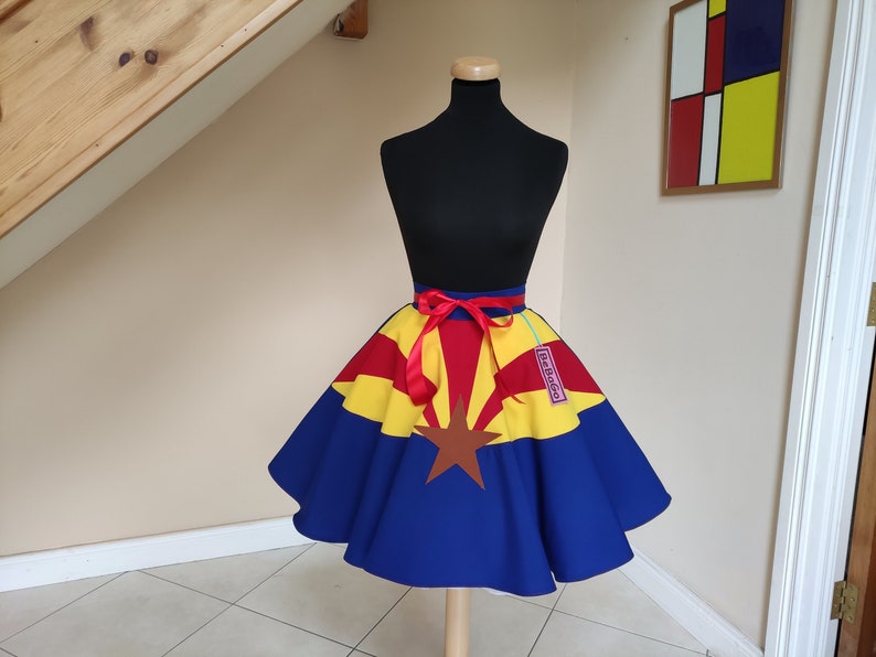 May include: A blue, yellow and red circle skirt with a red ribbon belt. The skirt is decorated with the Arizona state flag. The skirt is on a white mannequin.