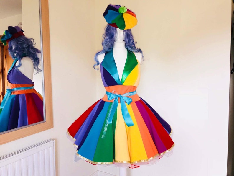 May include: A rainbow-colored dress with a matching hat. The dress has a fitted bodice and a full skirt. The hat is a round, flat-brimmed hat with a pom-pom on top.