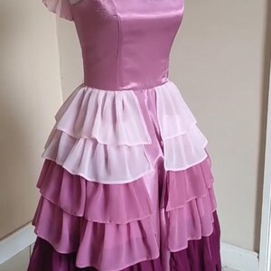 May include: A layered, floor-length dress in shades of pink and purple. The bodice is a solid, satin-like material, while the skirt features multiple tiers of ruffled chiffon. The dress is displayed on a mannequin.