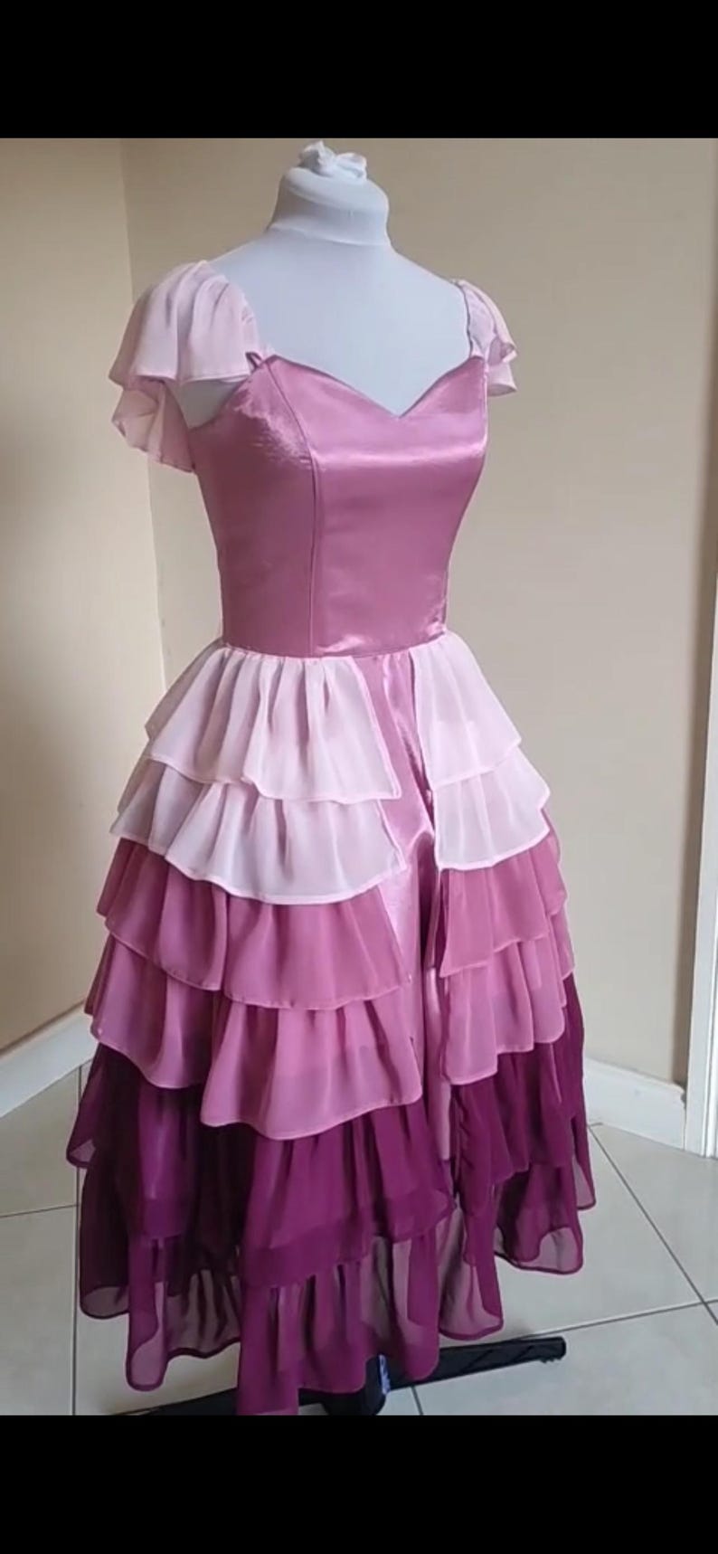 May include: A layered, floor-length dress in shades of pink and purple. The bodice is a solid, satin-like material, while the skirt features multiple tiers of ruffled chiffon. The dress is displayed on a mannequin.