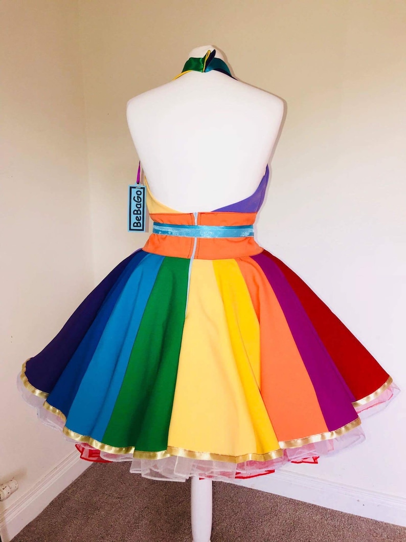 May include: A rainbow-colored dress with a halter neckline and a full skirt. The dress has a gold trim and a white petticoat. The dress is labeled "BeBoGo".