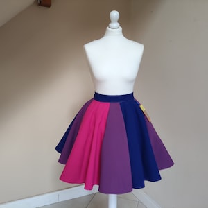 Può includere: Una gonna svasata nei colori della bandiera del pride bisessuale, esposta su un manichino bianco. La gonna presenta pannelli rosa, viola e blu, con una fascia in vita blu scuro. La gonna è un modello al ginocchio.