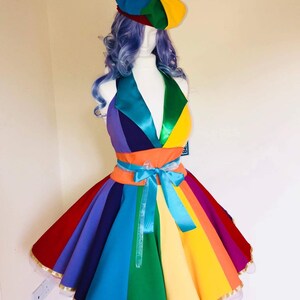 May include: A rainbow-colored dress with a matching beret. The dress has a fitted bodice and a full skirt with a rainbow stripe pattern. The skirt is trimmed with gold ribbon and has a blue ribbon tied around the waist.