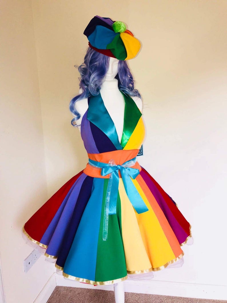 May include: A rainbow-colored dress with a matching beret. The dress has a fitted bodice and a full skirt with a rainbow stripe pattern. The skirt is trimmed with gold ribbon and has a blue ribbon tied around the waist.