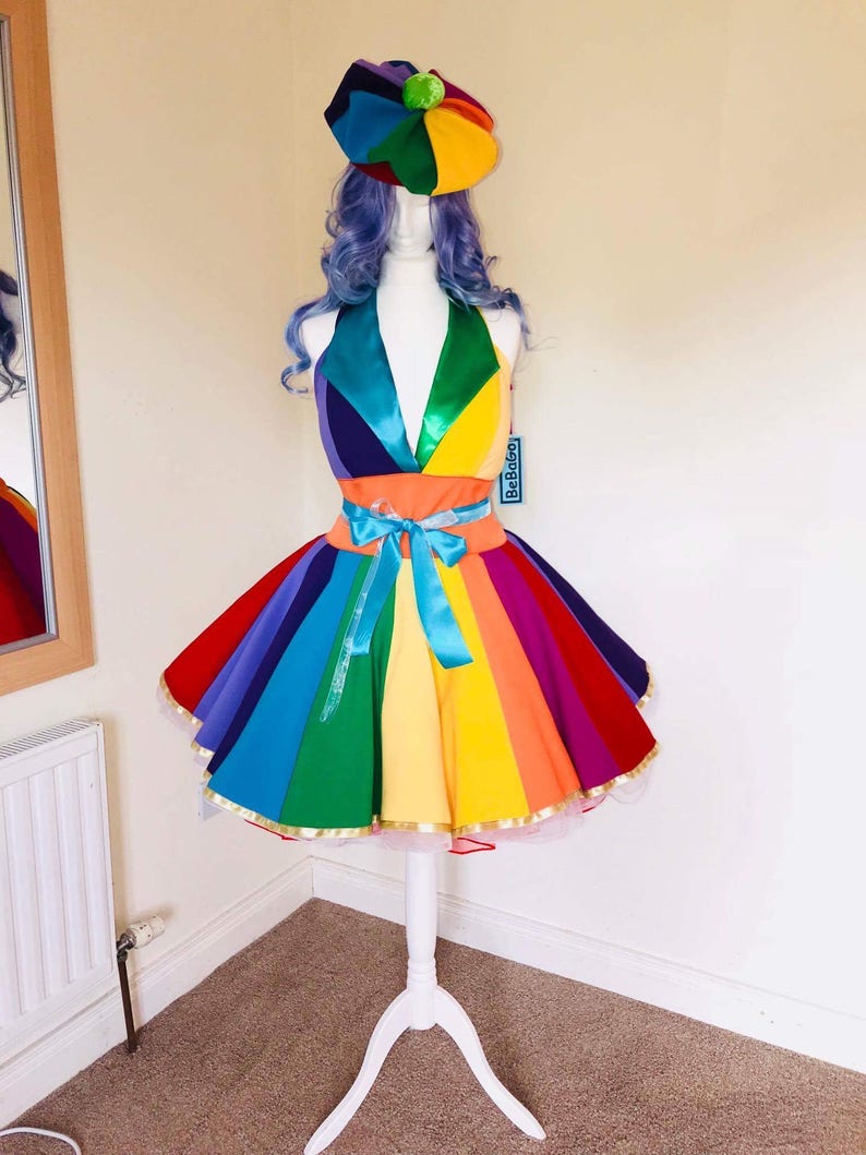 May include: A rainbow-colored dress with a matching hat. The dress has a fitted bodice and a full skirt with a tulle underskirt. The hat is a beret style with a pom-pom on top. The dress is on a white mannequin.