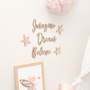 May include: Wooden wall decor featuring the words "Imagine Dream Believe" in cursive script, accompanied by four wooden stars. A framed print of a pink fairy with butterfly wings sits on a white shelf alongside a pink mushroom-shaped nightlight.