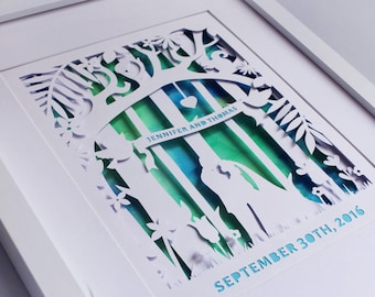 May include: A white paper cut art piece framed in white. The art depicts a couple standing under a tree with a banner that reads "Jennifer and Thomas" and the date "September 30th, 2016". The background is a watercolor wash of blue and green.