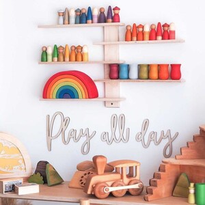 May include: A wooden rainbow arch with seven colors, wooden peg dolls in various colors, and a wooden shelf unit with the words "Play all day" on the wall.