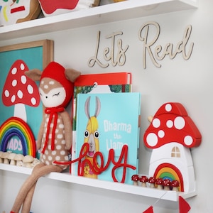 May include: A white shelf with a red and white mushroom toy, a rainbow toy, a stuffed deer toy, and a book titled "Dharma the Llama". The shelf also has a wooden sign that says "Let's Read" in cursive.