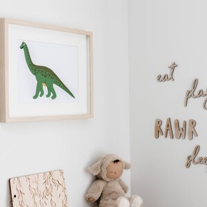 Op de afbeelding: Een ingelijste print van een groene dinosaurus met bruine vlekken op een witte achtergrond. De print hangt aan een witte muur. Er zijn houten letters op de muur die "eat", "play", "RAWR" en "sleep" spellen.