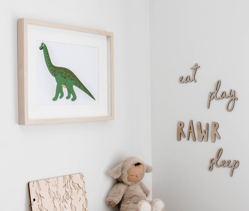 Op de afbeelding: Een ingelijste print van een groene dinosaurus met bruine vlekken op een witte achtergrond. De print hangt aan een witte muur. Er zijn houten letters op de muur die "eat", "play", "RAWR" en "sleep" spellen.